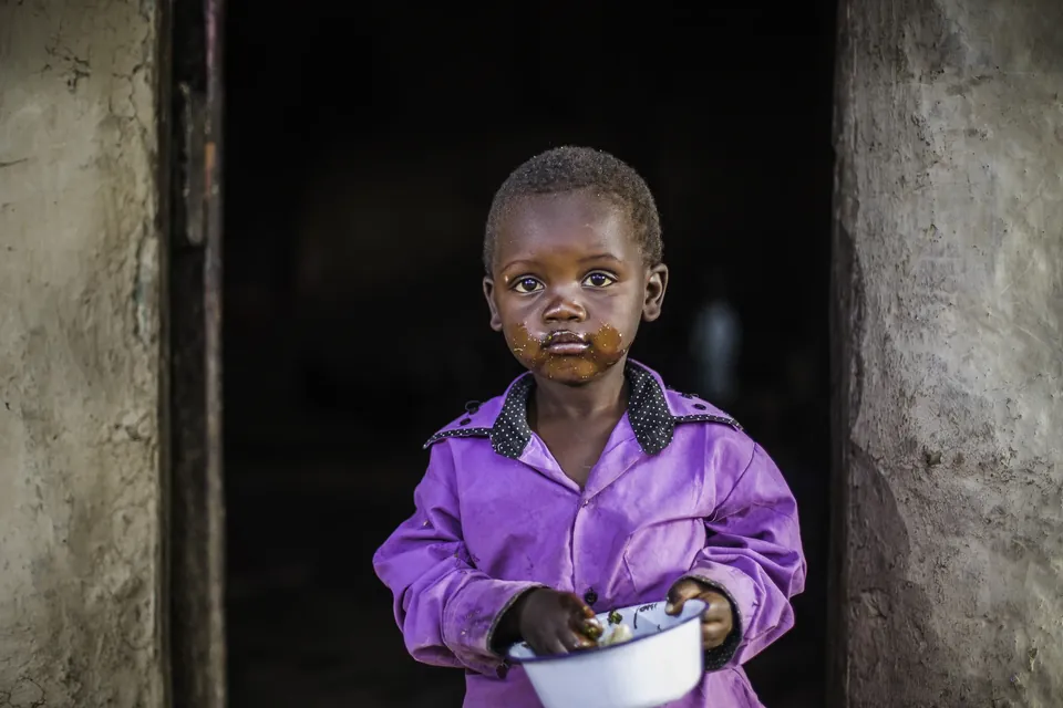 A small boy in a purple shirt holds a bowl in a doorway
