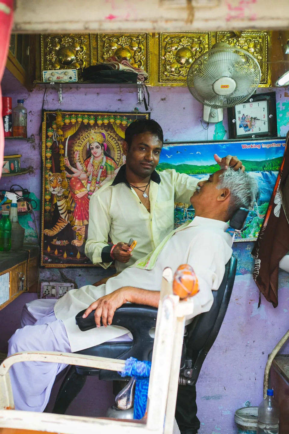 A barber shaves a customer in a small purple-walled shop in India